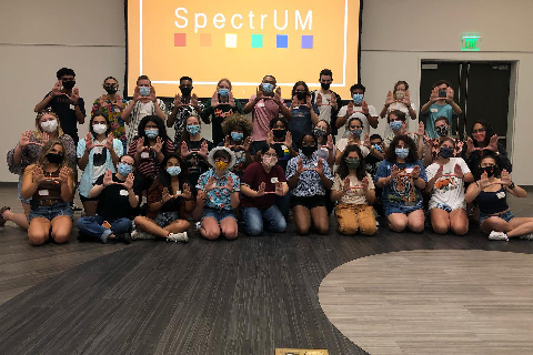 A group of SpectrUM students at an event stand together, holding up the U sign with their hands. They are all wearing face masks. 